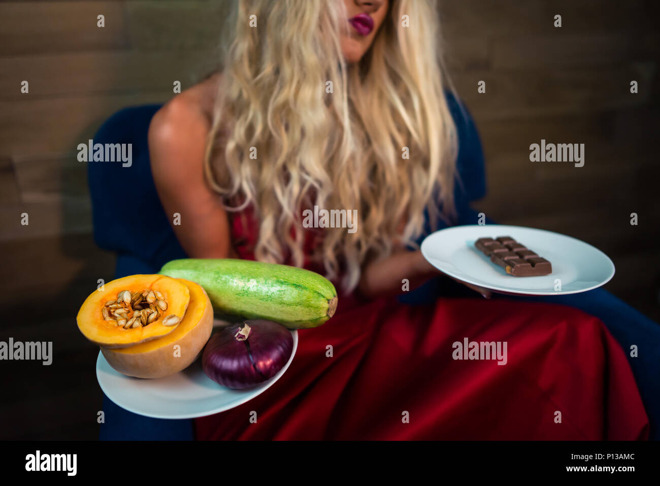 Giovane donna in abito rosso si siede sulla poltrona e trattiene il cibo in mani. Pretty girl scegliendo tra un sano o cibo malsano - Ortaggi o di cioccolato Foto Stock