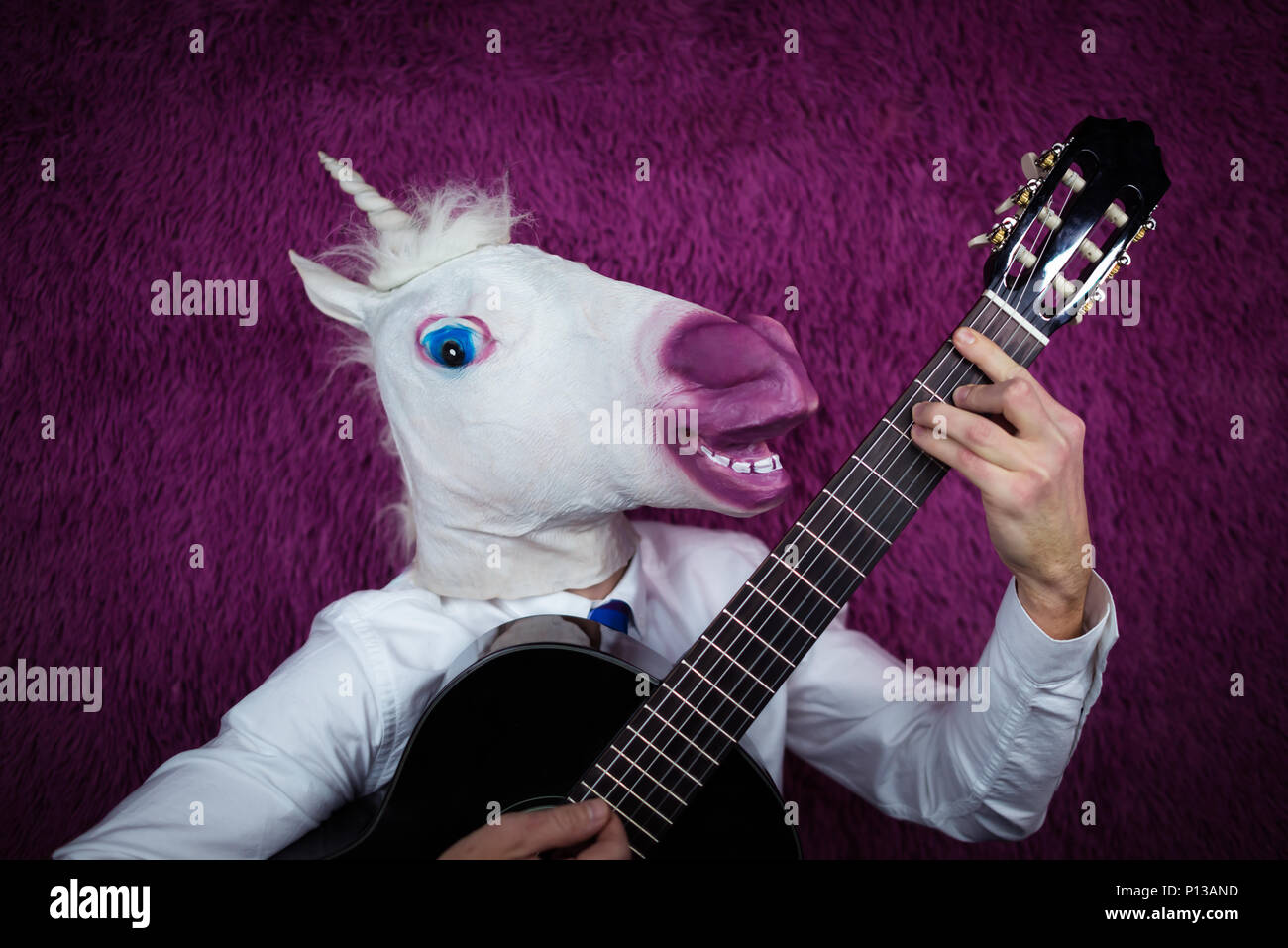 Freaky giovane uomo nella maschera di comico a suonare la chitarra su sfondo viola. Ritratto di un insolito ragazzo in camicia e cravatta con uno strumento musicale Foto Stock