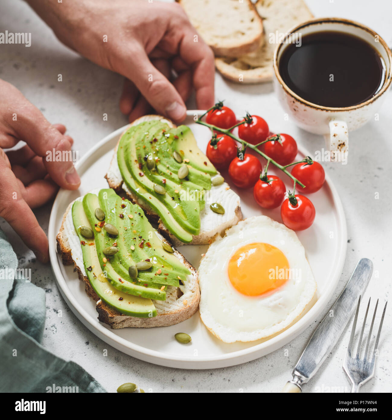 Toast con avocado, Uovo fritto, pomodori e tazza di caffè. Una sana prima colazione. Mani tenendo la piastra con una sana prima colazione il cibo. Concetto di vita sana Foto Stock