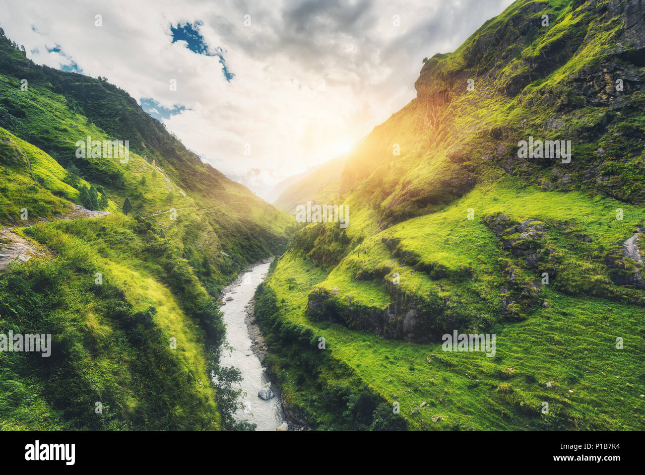 Vista con incredibili montagne ricoperte di erba verde, fiume, prati e bosco, cielo blu con nuvole e sole in autunno in Nepal al tramonto. Valle di montagna a Foto Stock