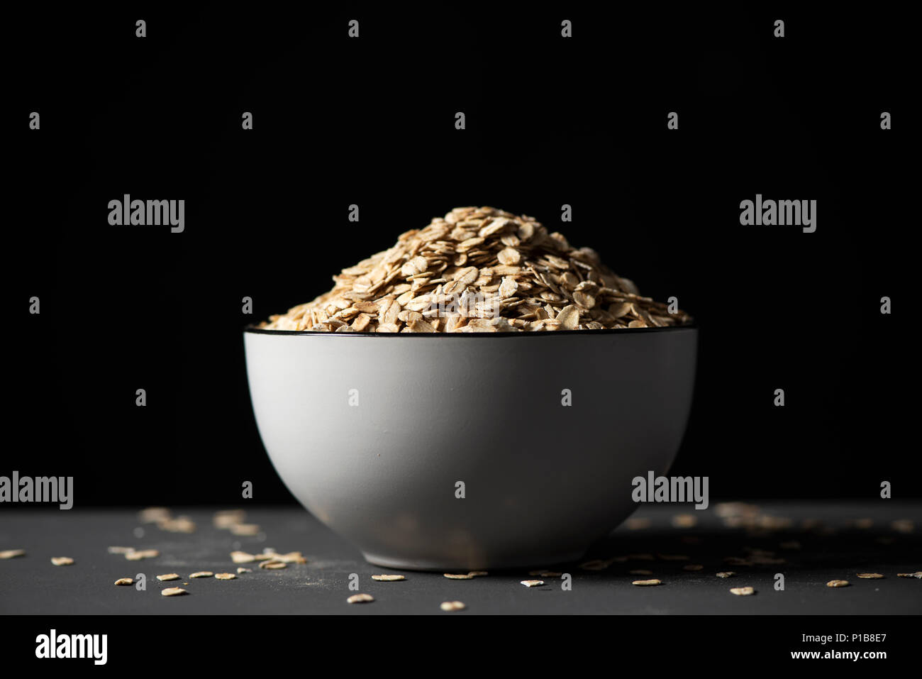 Primo piano di un bianco di ceramica ciotola piena di fiocchi d'avena su una tabella grigia, contro uno sfondo nero Foto Stock