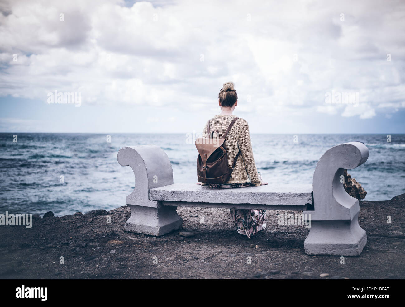 Vista posteriore della giovane donna in abiti lunghi e con zaino seduta sul banco di pietra dal mare Foto Stock
