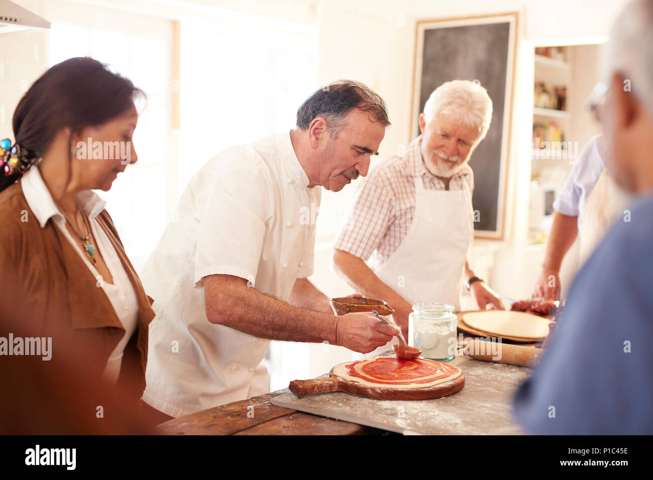 Senior gli amici a guardare lo chef diffusione di salsa marinara sulla pasta della pizza nella classe di cucina Foto Stock