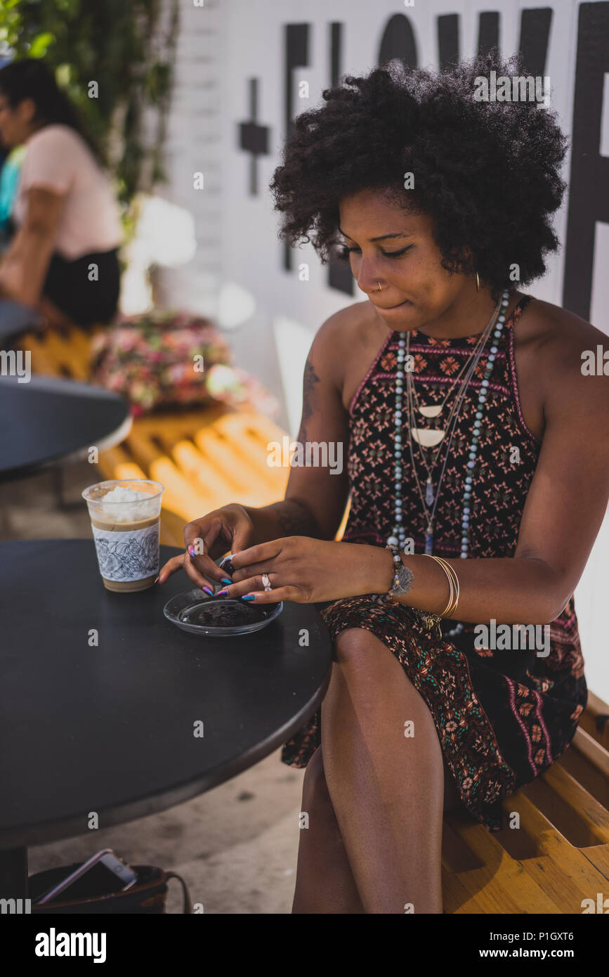 Donna nera ciambella di mangiare e di bere il caffè al di fuori in un coffee shop Foto Stock