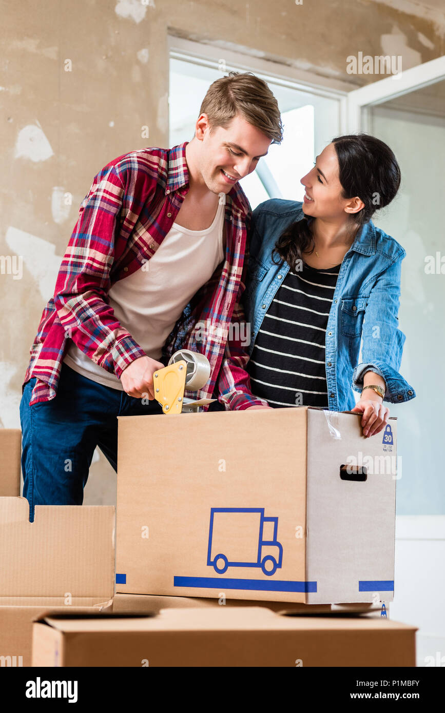 Il giovane e la sua ragazza la tenuta di una brown scatola di cartone con nastro adesivo mentre rinnovare insieme la loro nuova casa Foto Stock