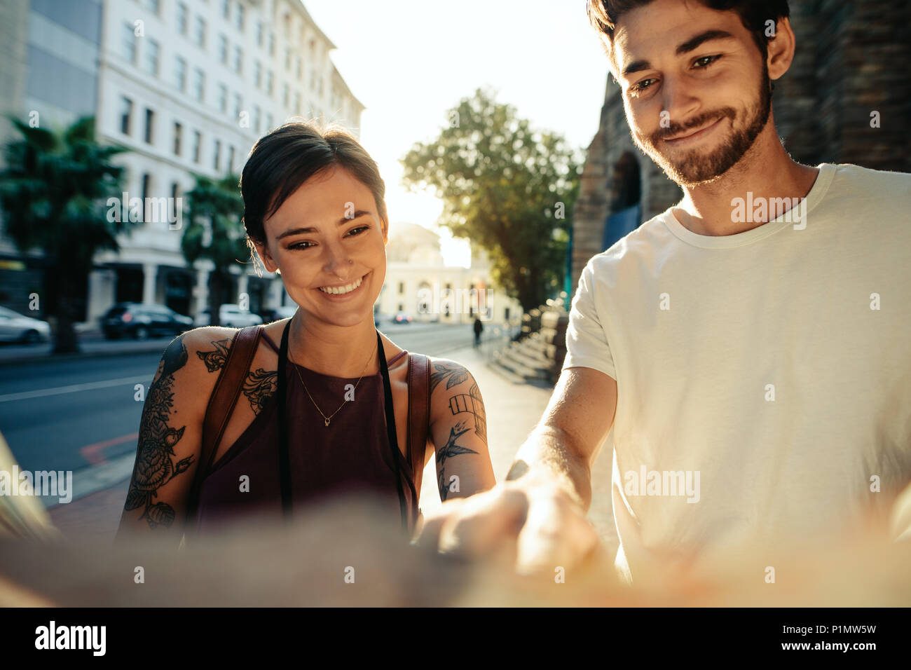 Turista giovane utilizzando una mappa per esplorare la città. Uomo sorridente e donna in piedi da una strada guardando la mappa con Sun in background. Foto Stock