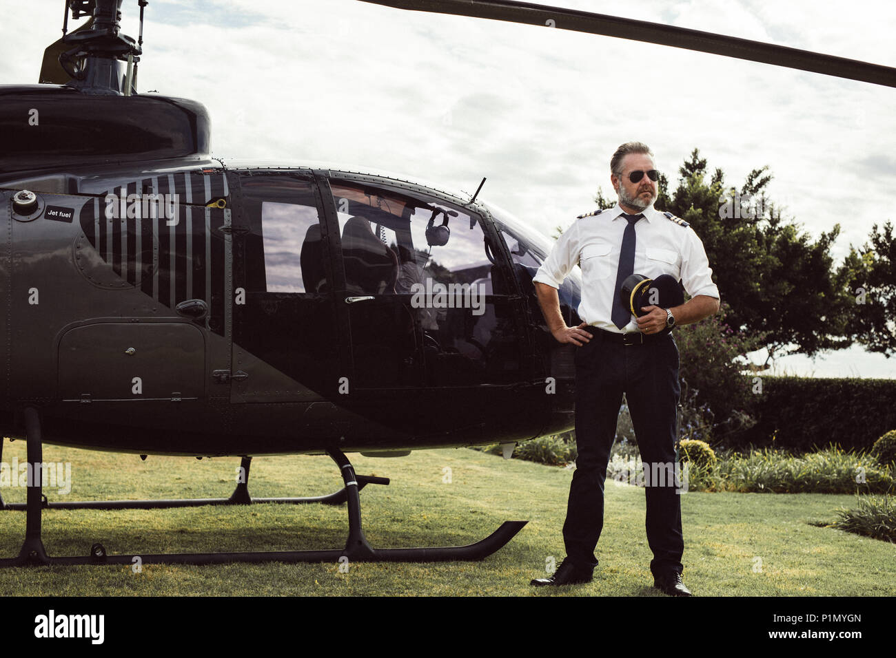 Maschio maturo pilota in uniforme al di fuori in piedi con un elicottero privato. Elicottero privato parcheggiato nel prato con pilota maschio in attesa. Foto Stock