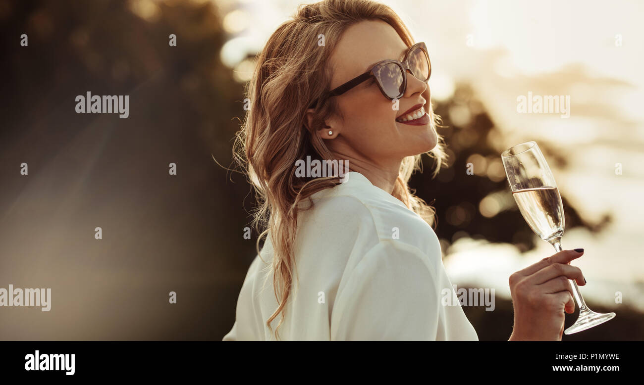 Donna sorridente in occhiali da sole di bere vino. Bellissimo modello femminile con un bicchiere di vino all'esterno. Foto Stock