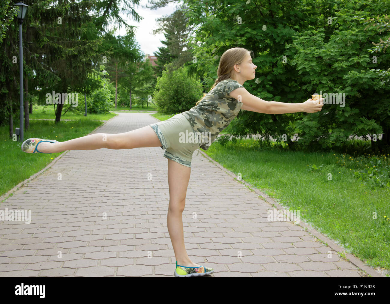 Slim ragazza adolescente con gelato nelle mani di danza nel parco godendo le prossime vacanze. Esercizio coreografico di bilanciamento su un piede su un ave Foto Stock