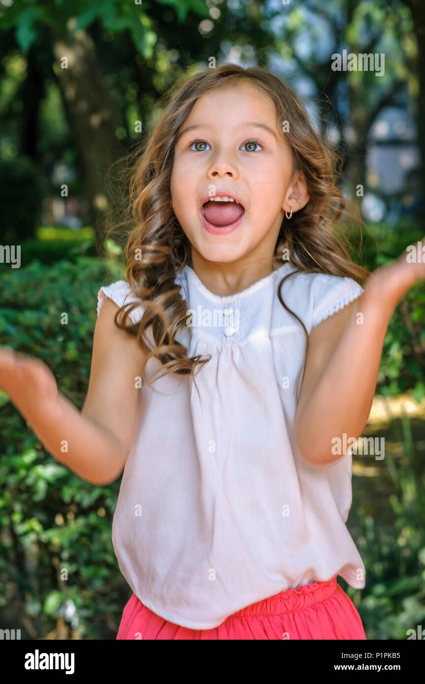 Carino cinque anni bambina con gli occhi blu e i capelli biondi che mostra la felicità e la sorpresa sul suo viso con un parco come sfondo Foto Stock