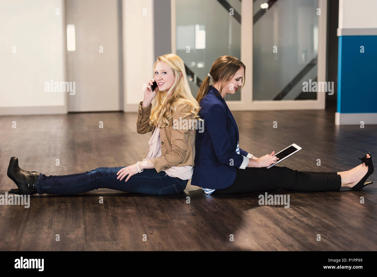 Due belle giovani donne che sono la millenaria professionisti seduti di schiena sul pavimento del proprio ambiente di lavoro utilizzando la tecnologia Foto Stock