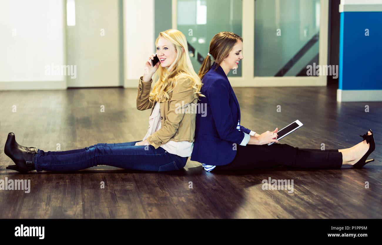 Due belle giovani donne che sono la millenaria professionisti seduti di schiena sul pavimento del proprio ambiente di lavoro utilizzando la tecnologia Foto Stock