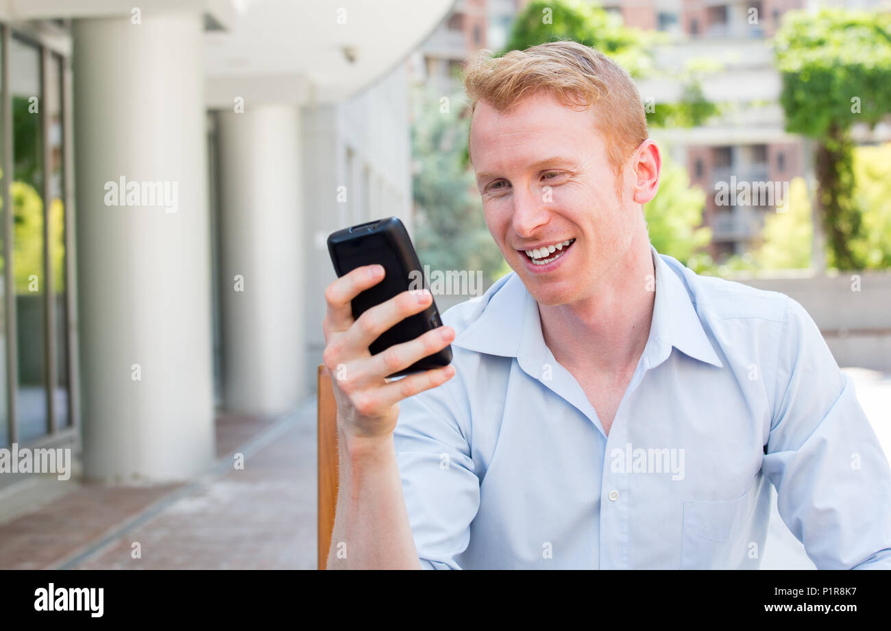 Closeup ritratto, giovani happy businessman in maglietta blu seduta, controllando il suo cellulare, isolato sullo sfondo di un edificio cittadino, alberi, su un soleggiato Foto Stock