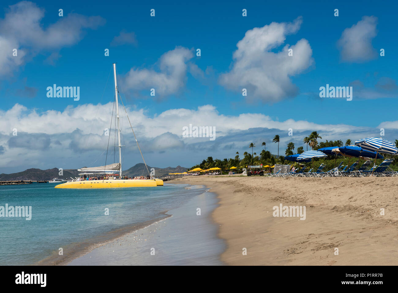Il catamarano giallo e ombrelli a strisce lungo la spiaggia di Isola dei Caraibi; Nevis, West Indies Foto Stock