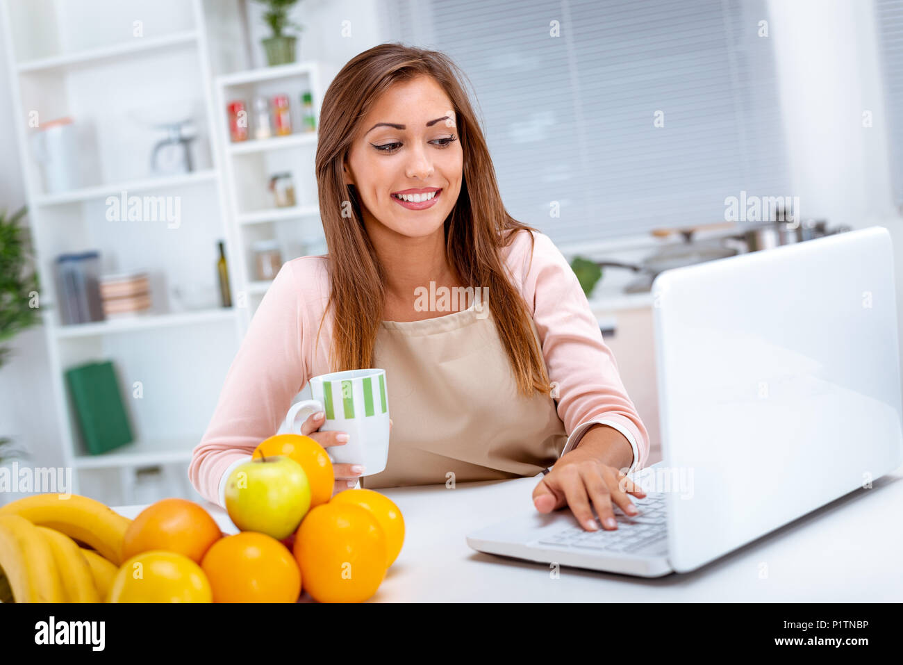 Bella giovane donna a bere caffè e la ricetta di lettura su laptop in cucina. Foto Stock