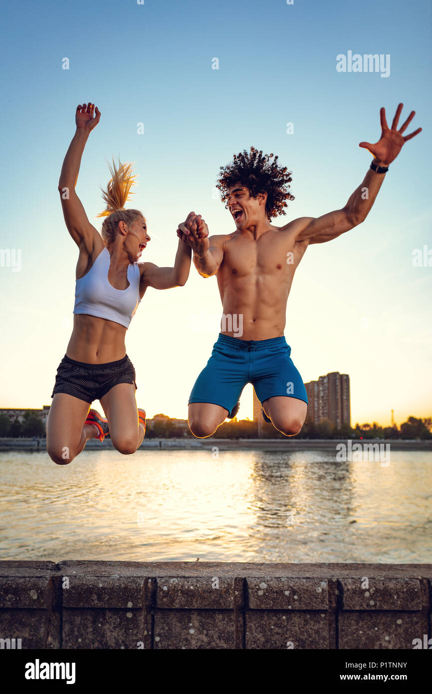 Giovane e bella coppia sorridente e saltando dopo il successo formativo dal corso del fiume al tramonto. Foto Stock