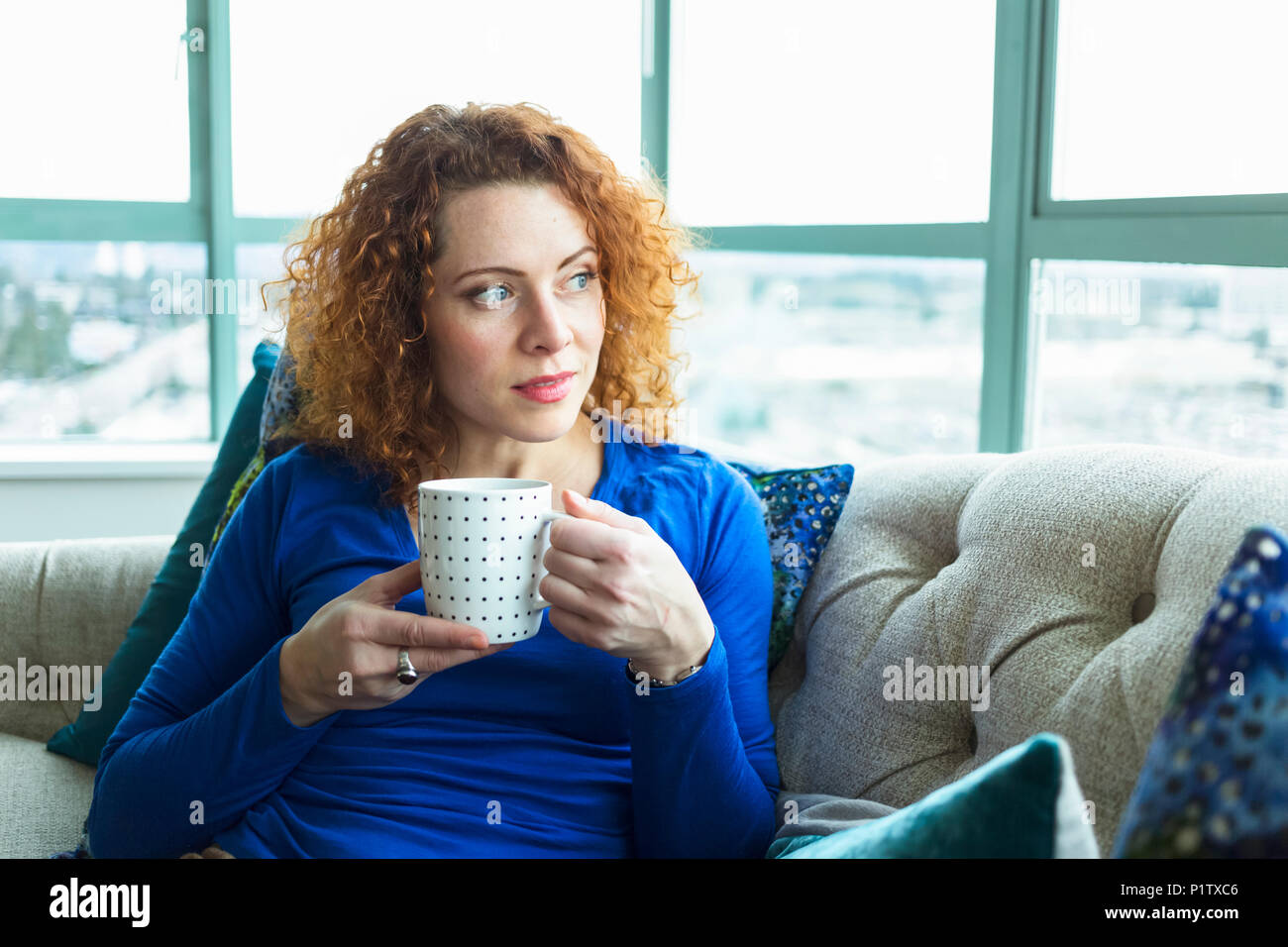 Donna con rosso, capelli ricci seduta con una tazza da una finestra; Surrey, British Columbia, Canada Foto Stock