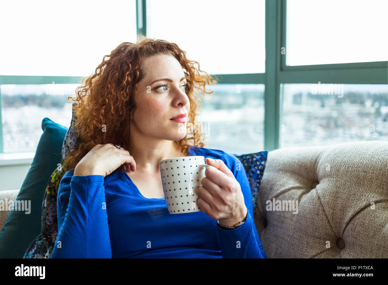 Donna con rosso, capelli ricci seduta con una tazza guardando fuori dalla finestra; Surrey, British Columbia, Canada Foto Stock