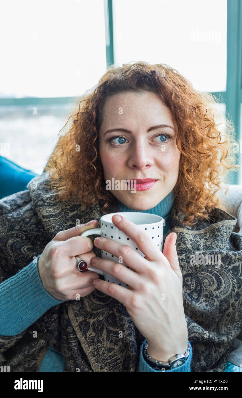 Donna con rosso, capelli ricci seduta con una tazza da una finestra; Surrey, British Columbia, Canada Foto Stock