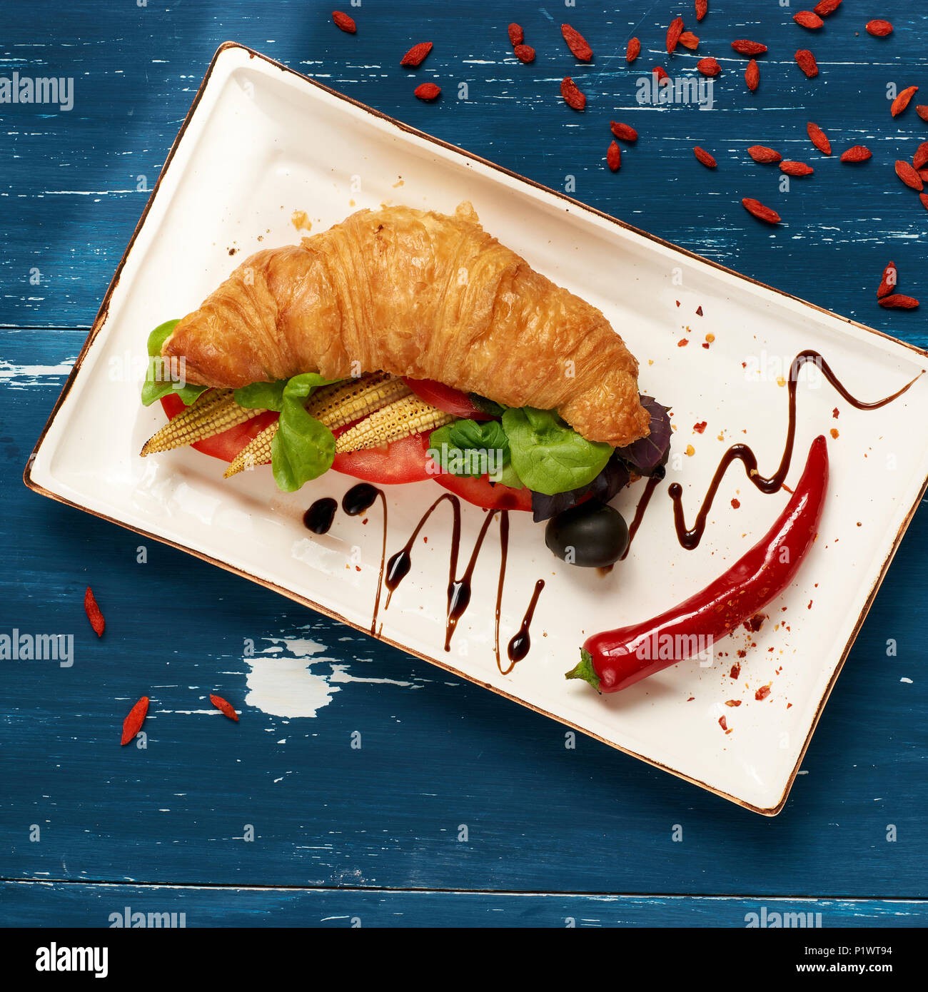 Appetitosi sandwich vegetariano con croissant e verdure giacente sulla piastra piana di forma rettangolare, ciotola di olive e pomodori ciliegia ed essiccati goji bacche sc Foto Stock
