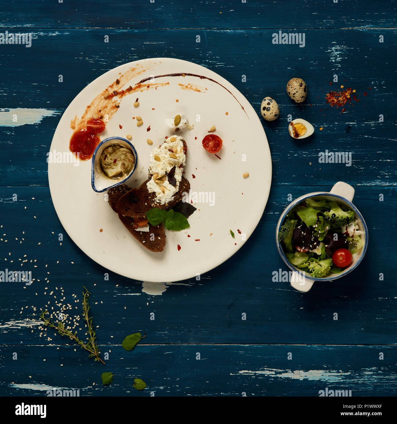 La bruschetta o le fette di pane condito con la crema di formaggio che giace sulla piastra rotonda e le verdure in piccoli pot permanente blu sulla superficie di legno. Vegetari sani Foto Stock