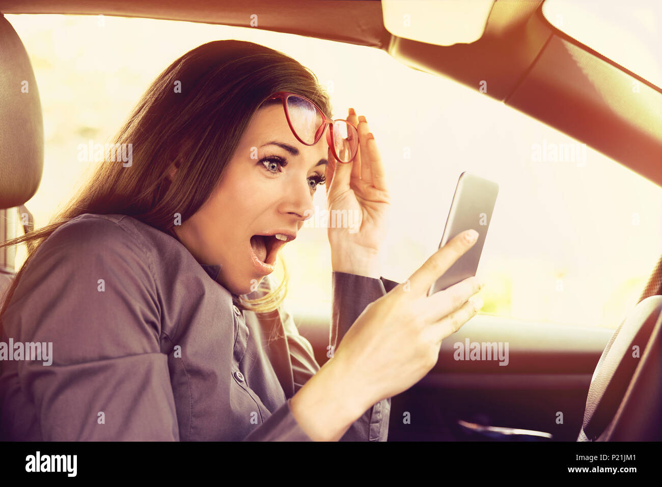 Giovane donna distratti la lettura di un messaggio sul cellulare, stupito, mentre si è alla guida di un veicolo Foto Stock