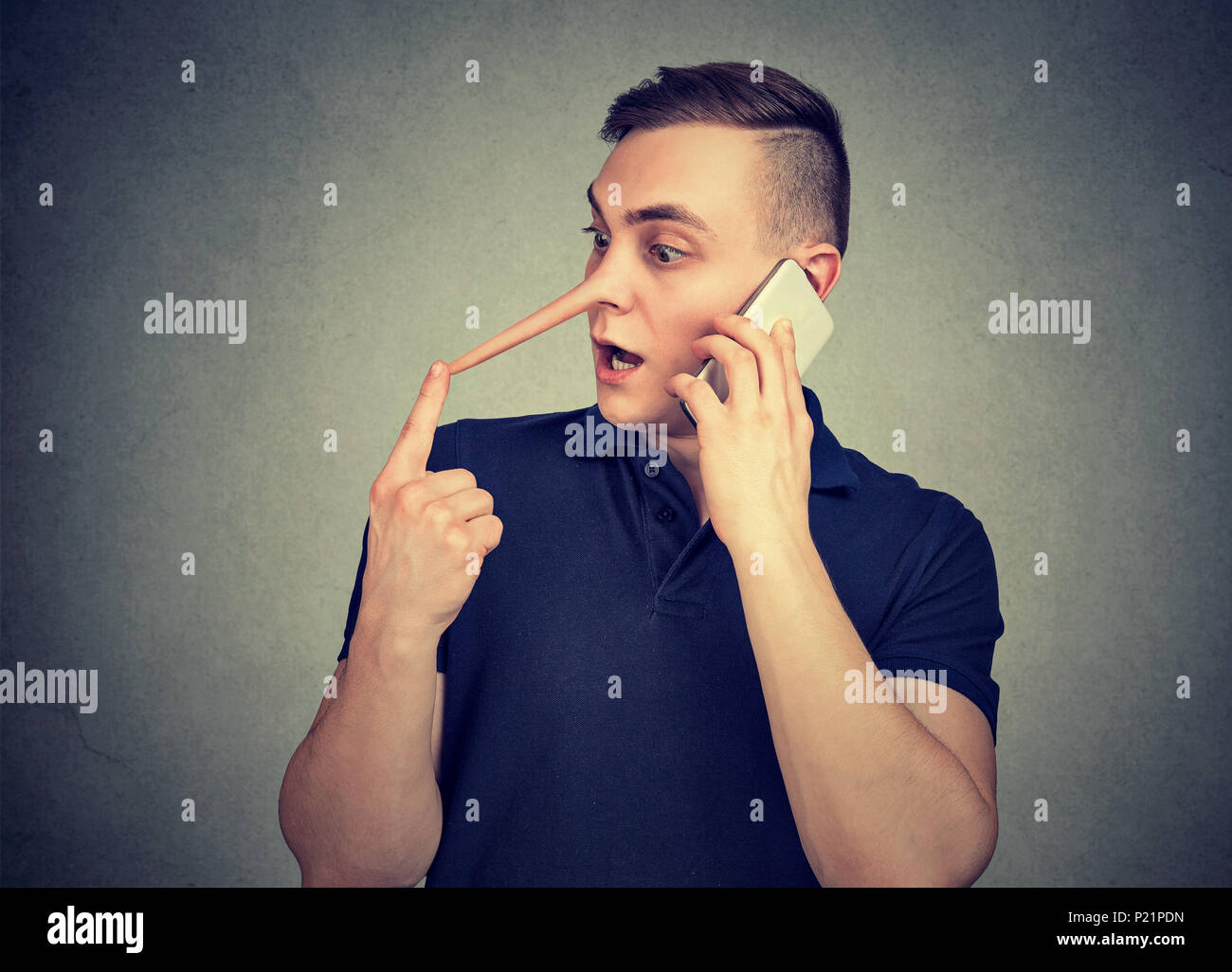 Giovane uomo hypocritic con il naso lungo parlando su smartphone e giacente espressamente su sfondo grigio. Foto Stock