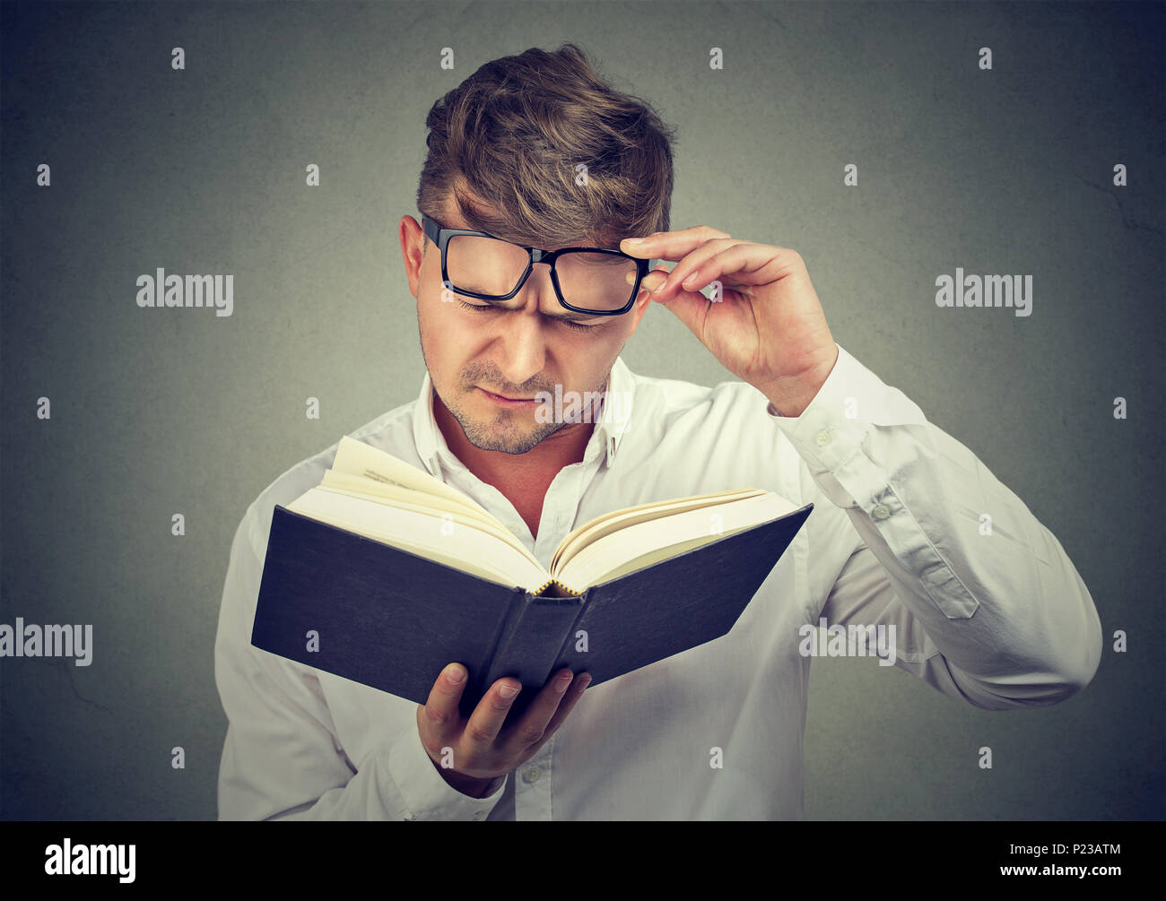 Giovane uomo accigliata sopracciglia durante il sollevamento di occhiali e cercando di leggere il libro su sfondo grigio. Foto Stock
