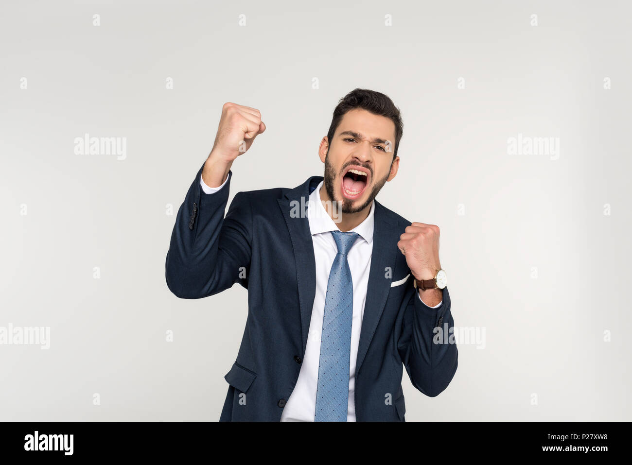 Eccitato giovane impreditore agitando i pugni e urlando in telecamera isolato su grigio Foto Stock