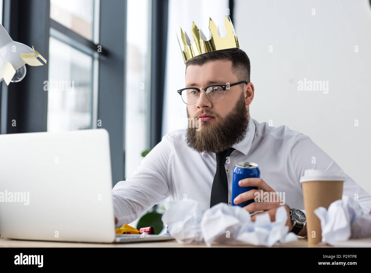 Ritratto di imprenditore con corona di carta sulla testa e soda drink in mano lavorando sul computer portatile al lavoro in ufficio Foto Stock