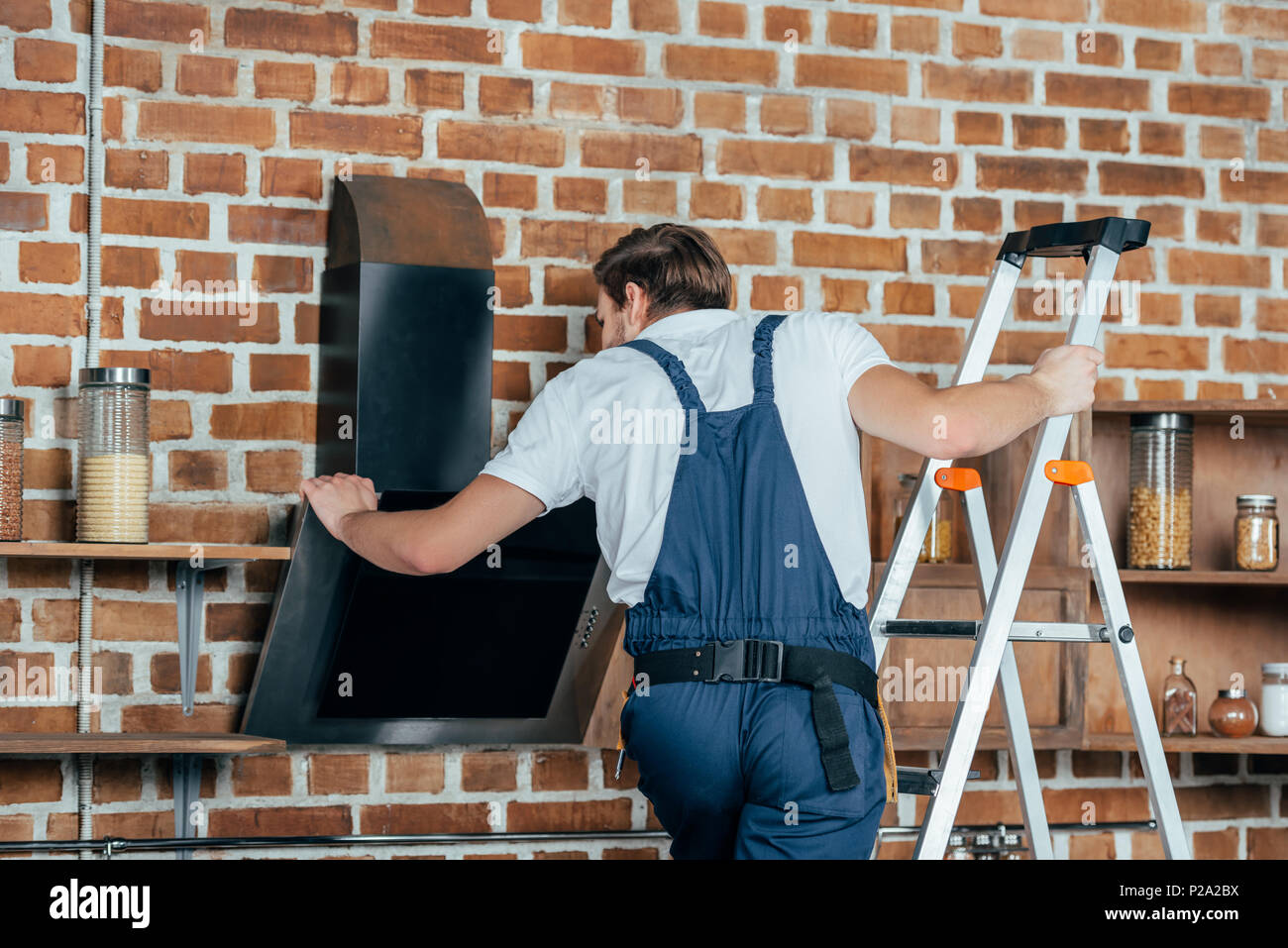 Giovane maestro in indumenti da lavoro protettiva permanente sulla scaletta e la riparazione di cappa da cucina Foto Stock