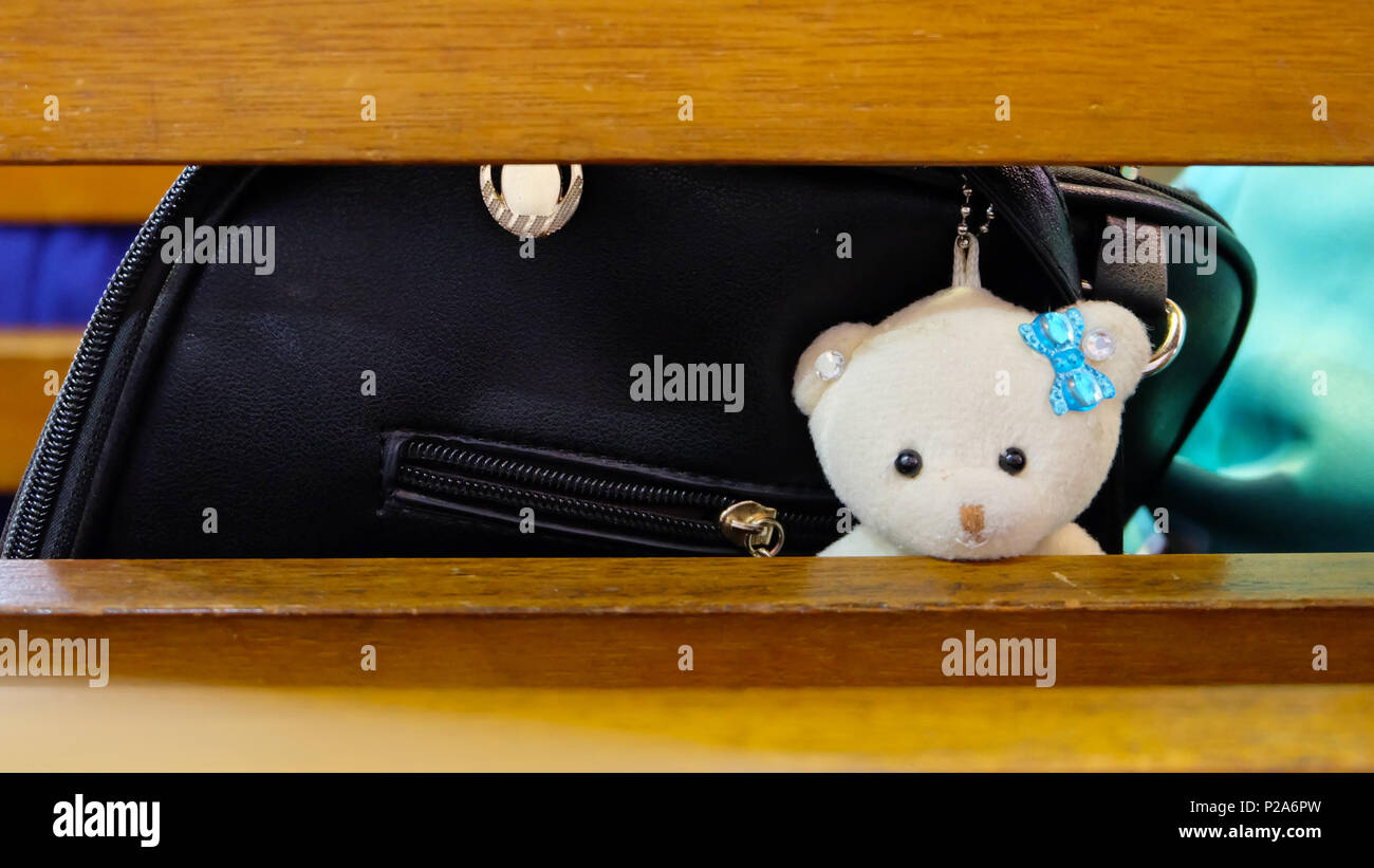 Una crema leggera bear lussureggiante con nastro blu appeso ad una borsa nera del peering attraverso lo schienale di una panca in legno Foto Stock