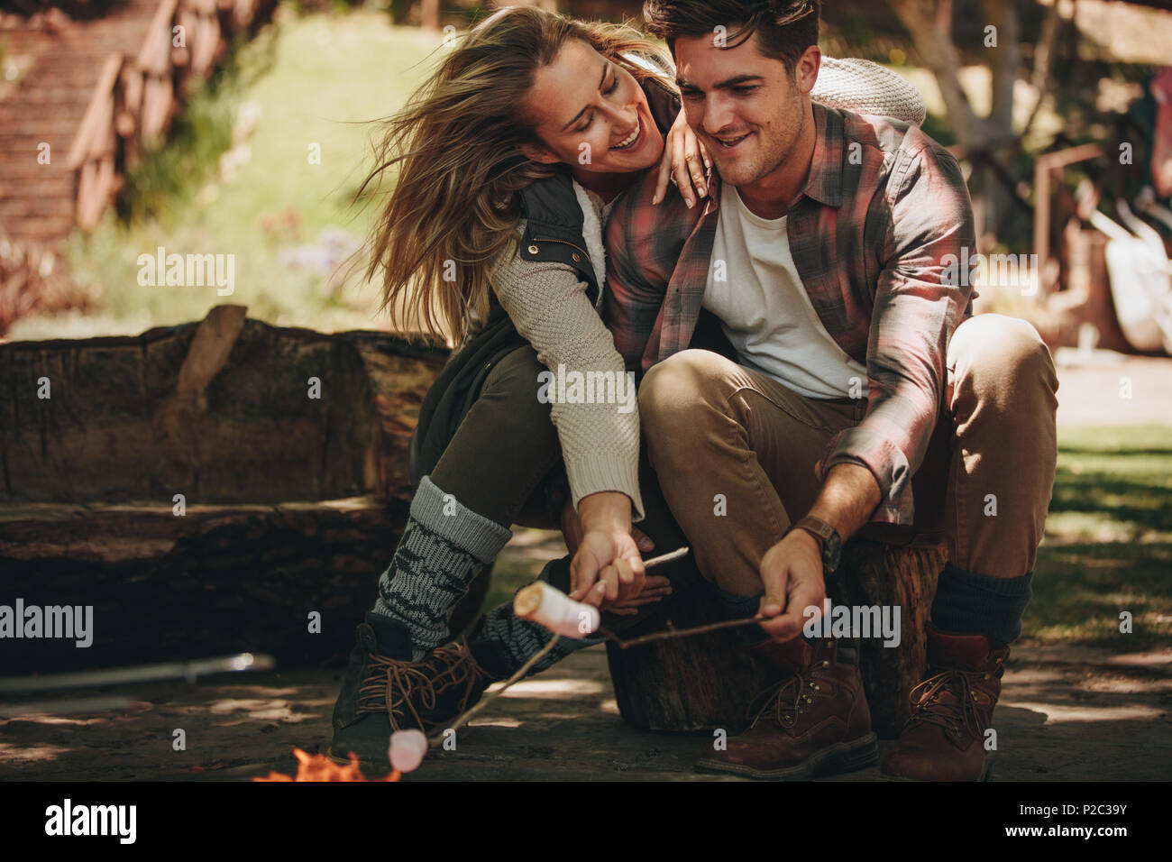 Felice l'uomo e donna seduta dal fuoco ed arrostire marshmallows su bastoni al campeggio. Coppia romantica avente un grande tempo sul loro camping. Foto Stock