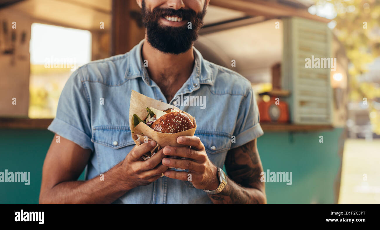 Chiusura del giovane uomo mangiare hamburger all'esterno. Uomo con barba avente street food. Foto Stock