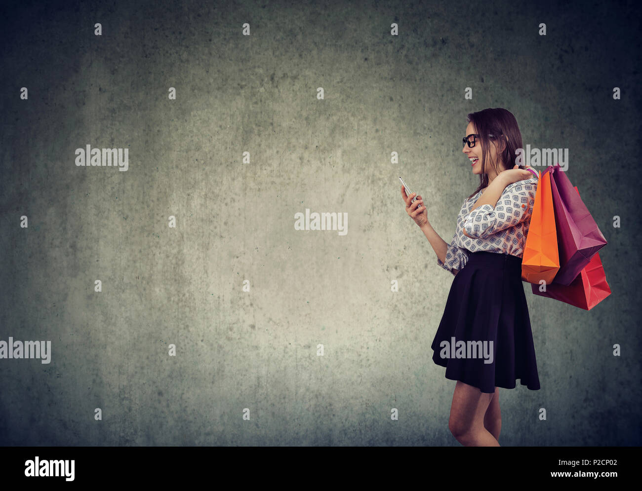 Vista laterale del casual ragazza shopping via permanente dello smartphone con abbondanza di sacchetti di carta e sorridente felicemente. Foto Stock