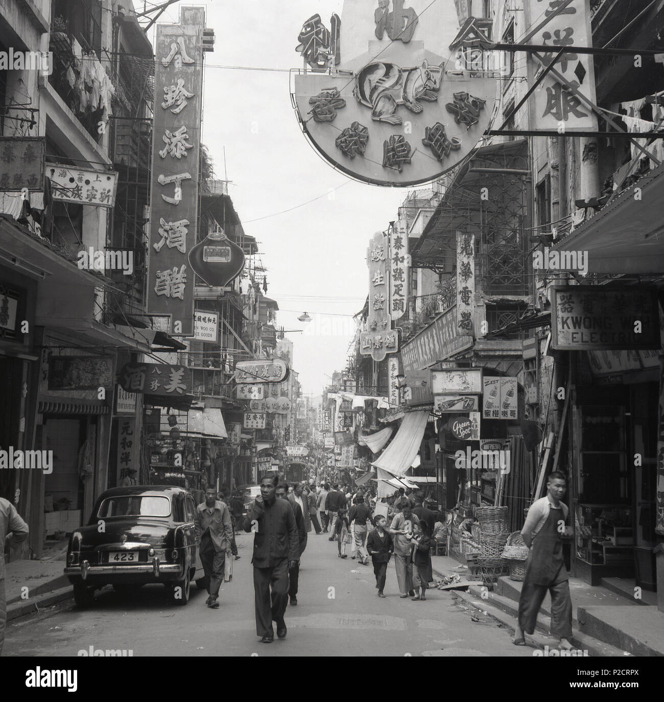 Anni '50, storico, una foto di J Allan Cash da questa epoca guardando giù una strada nella città vecchia di Hong Kong, Asia, mostrando la gente che cammina per la strada, negozi, insegne cinesi e l'architettura della zona, con la sistemazione vivente sopra i punti vendita al dettaglio. Anche nella foto, parcheggiato in strada, un'auto dell'epoca con targa, 423. Foto Stock