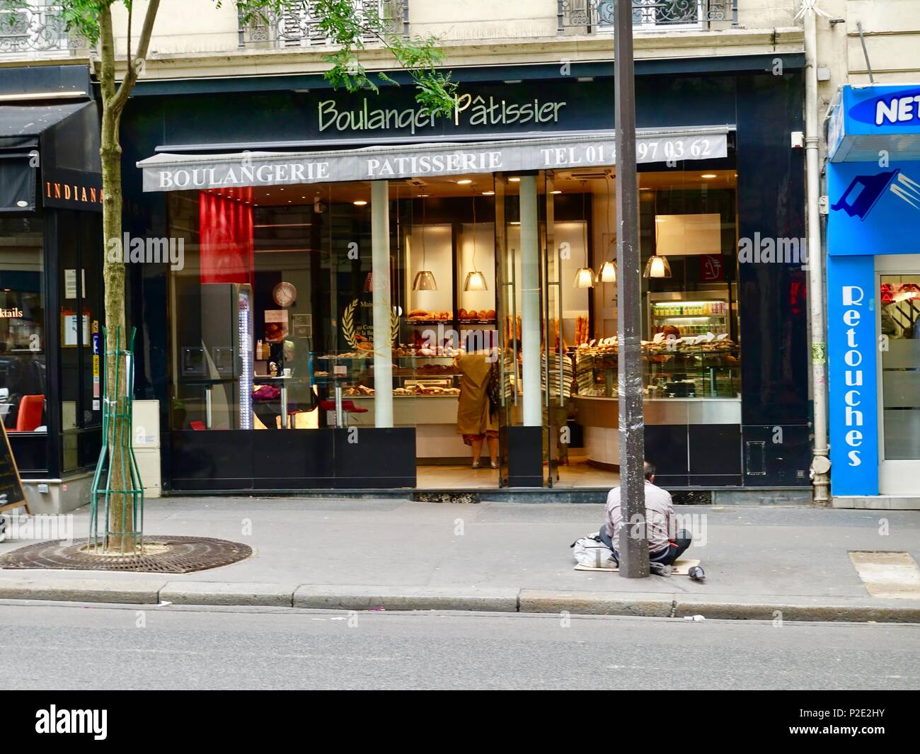 Senzatetto uomo seduto sul marciapiede al di fuori di un panificio mentre donna ben vestito donna negozi dentro, Parigi, Francia Foto Stock
