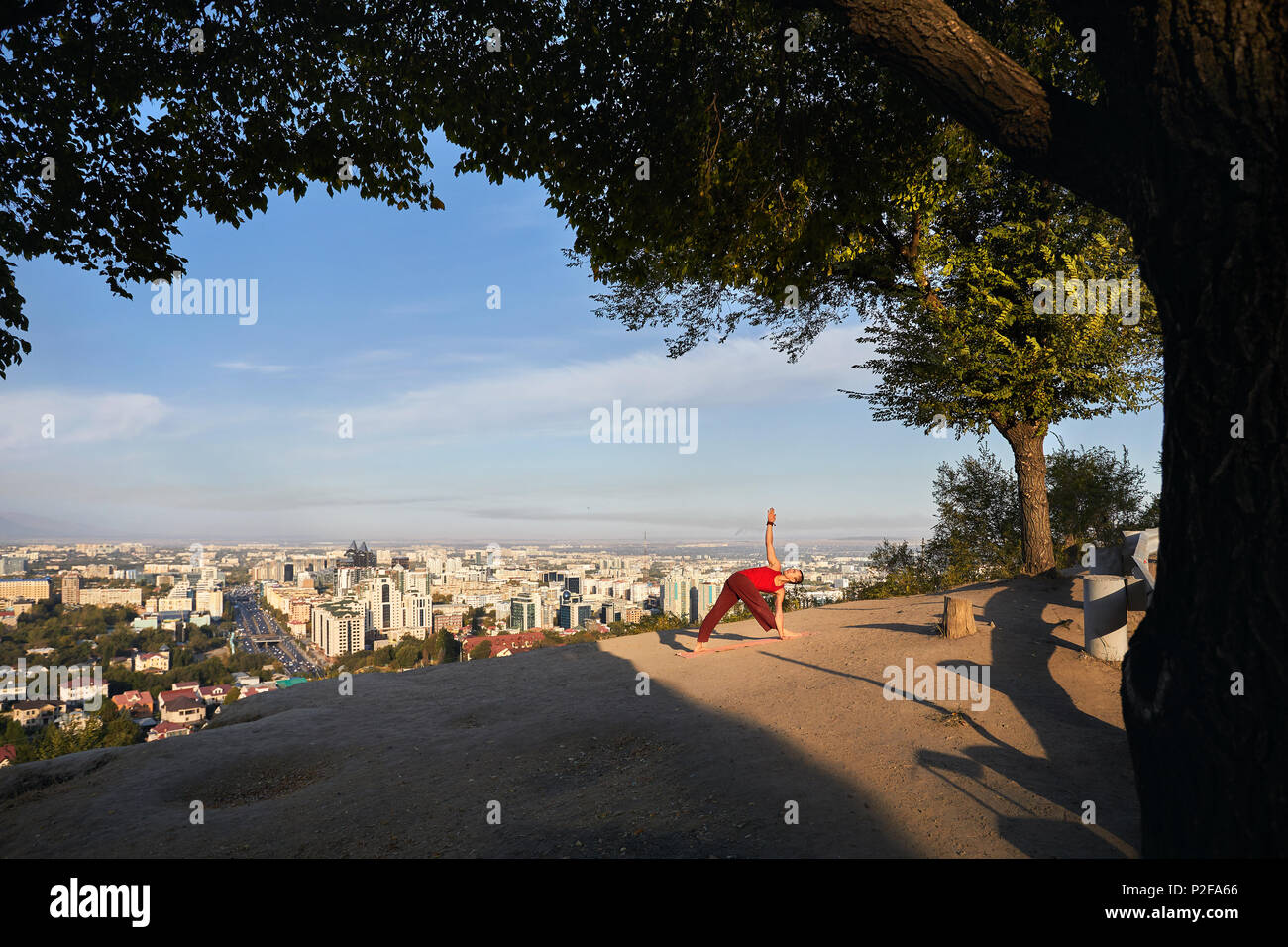 Montare l'uomo in costume rosso facendo il triangolo di Yoga asana nel parco con città e montagna in background in Almaty, Kazakhstan Foto Stock
