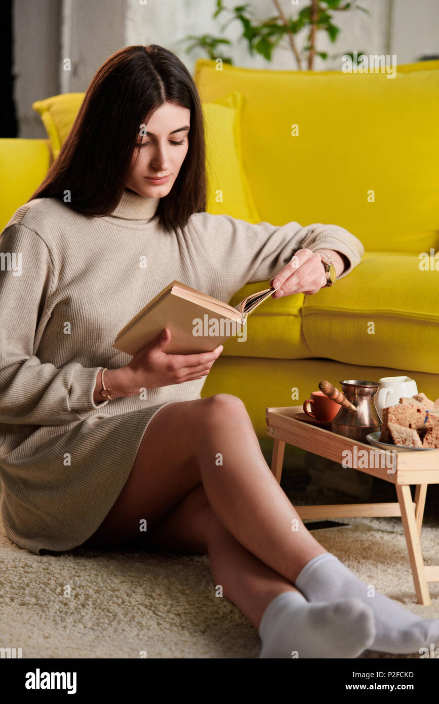 Donna libro lettura mentre è seduto sul pavimento vicino al vassoio in legno con la tazza di caffè e pasticceria a casa Foto Stock