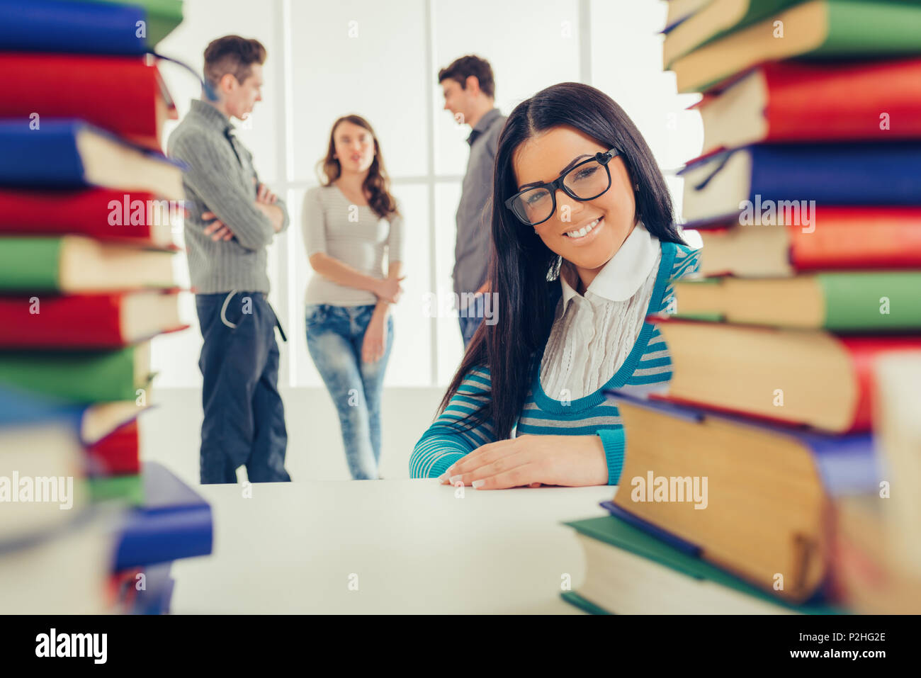 Ritratto di una bella ragazza sorridente con gli occhiali seduto dietro i molti libri in primo piano. Guardando alla fotocamera. Un suo amici è permanente behin Foto Stock