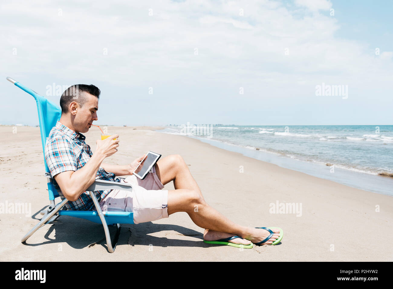 Primo piano di un giovane uomo caucasico, seduti su una sedia a sdraio sulla spiaggia, utilizzando una tavoletta digitale mentre si beve una fresca bevanda a base di arancia Foto Stock