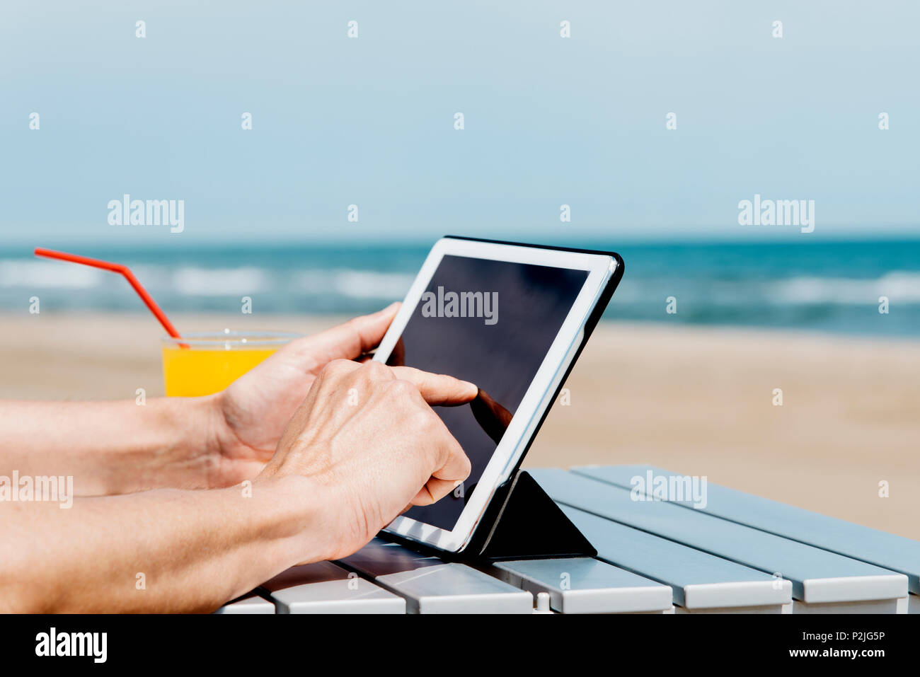 Primo piano di un giovane uomo caucasico sulla spiaggia, utilizzando una tavoletta digitale, seduti a un tavolo all'aperto, accanto a un bicchiere con una fresca bevanda a base di arancia Foto Stock