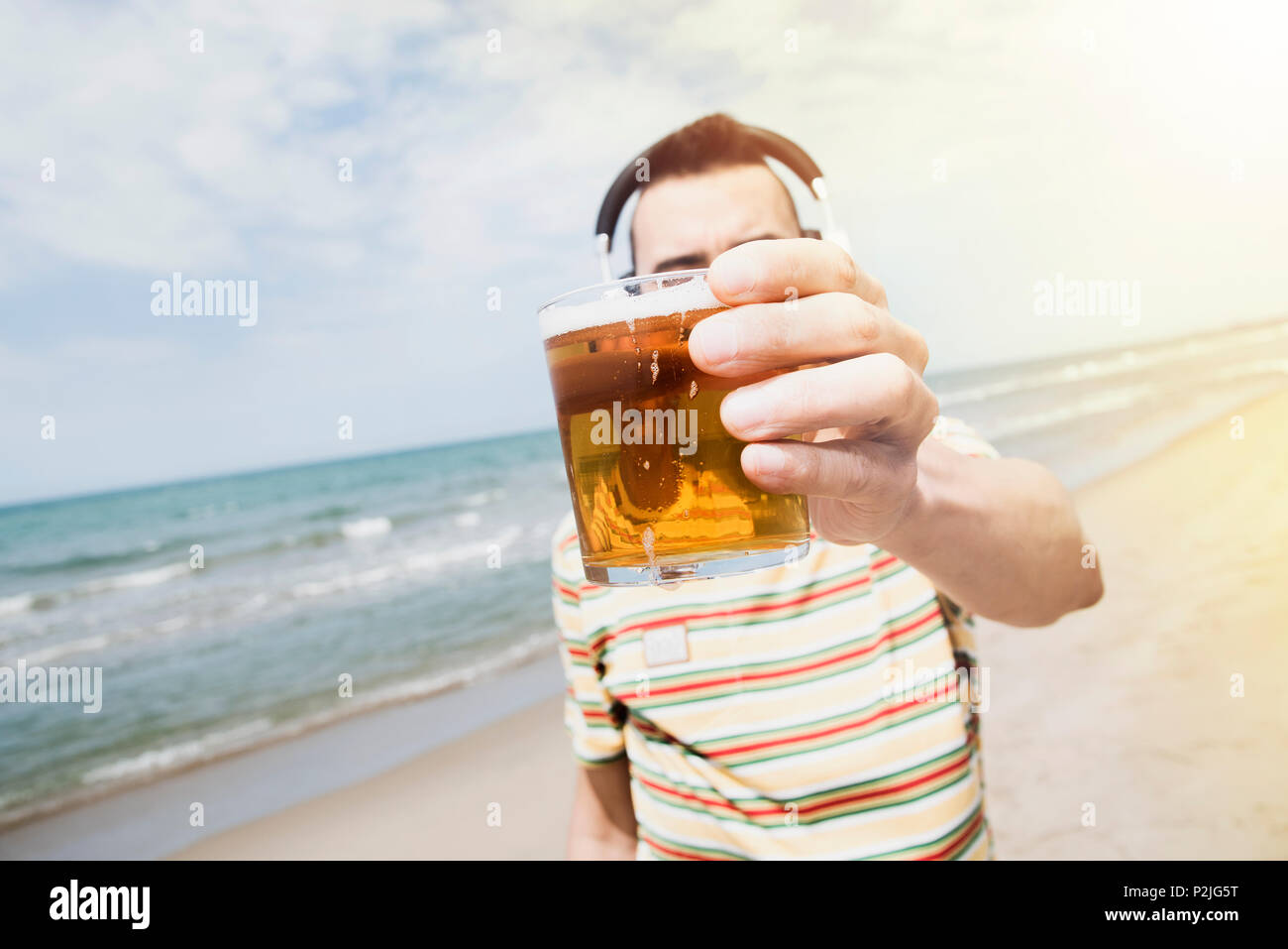 Primo piano di un giovane uomo caucasico sulla spiaggia ascoltando la musica con una coppia di cuffie senza fili mentre sta bevendo una birra di fronte al mare, con Foto Stock
