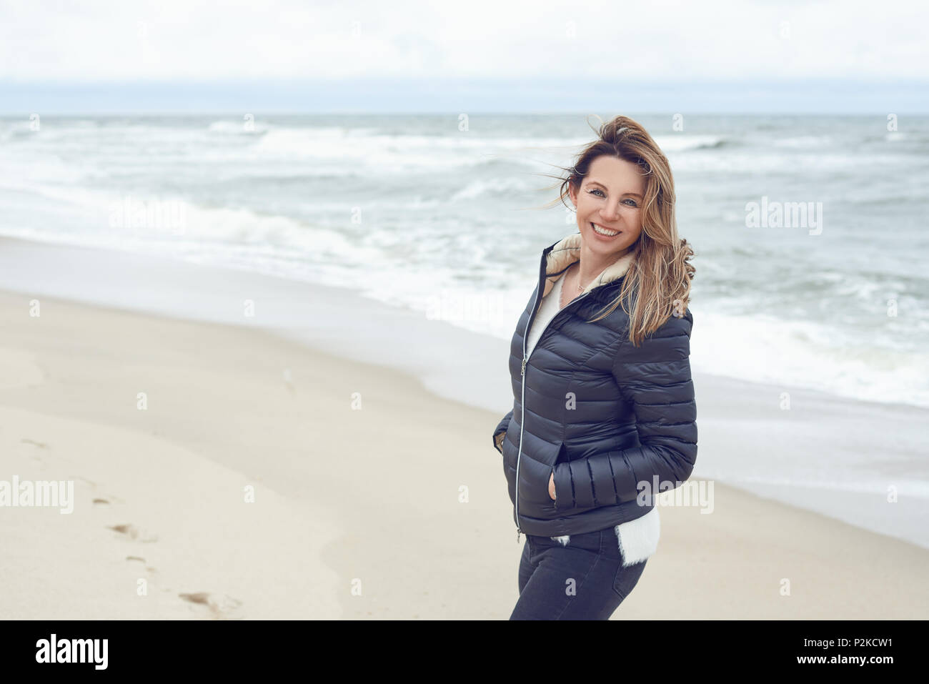 Attraente donna alla moda a camminare su una deserta spiaggia di sabbia in un giorno nuvoloso con la bassa marea girando di sorridere alla telecamera con spazio di copia Foto Stock