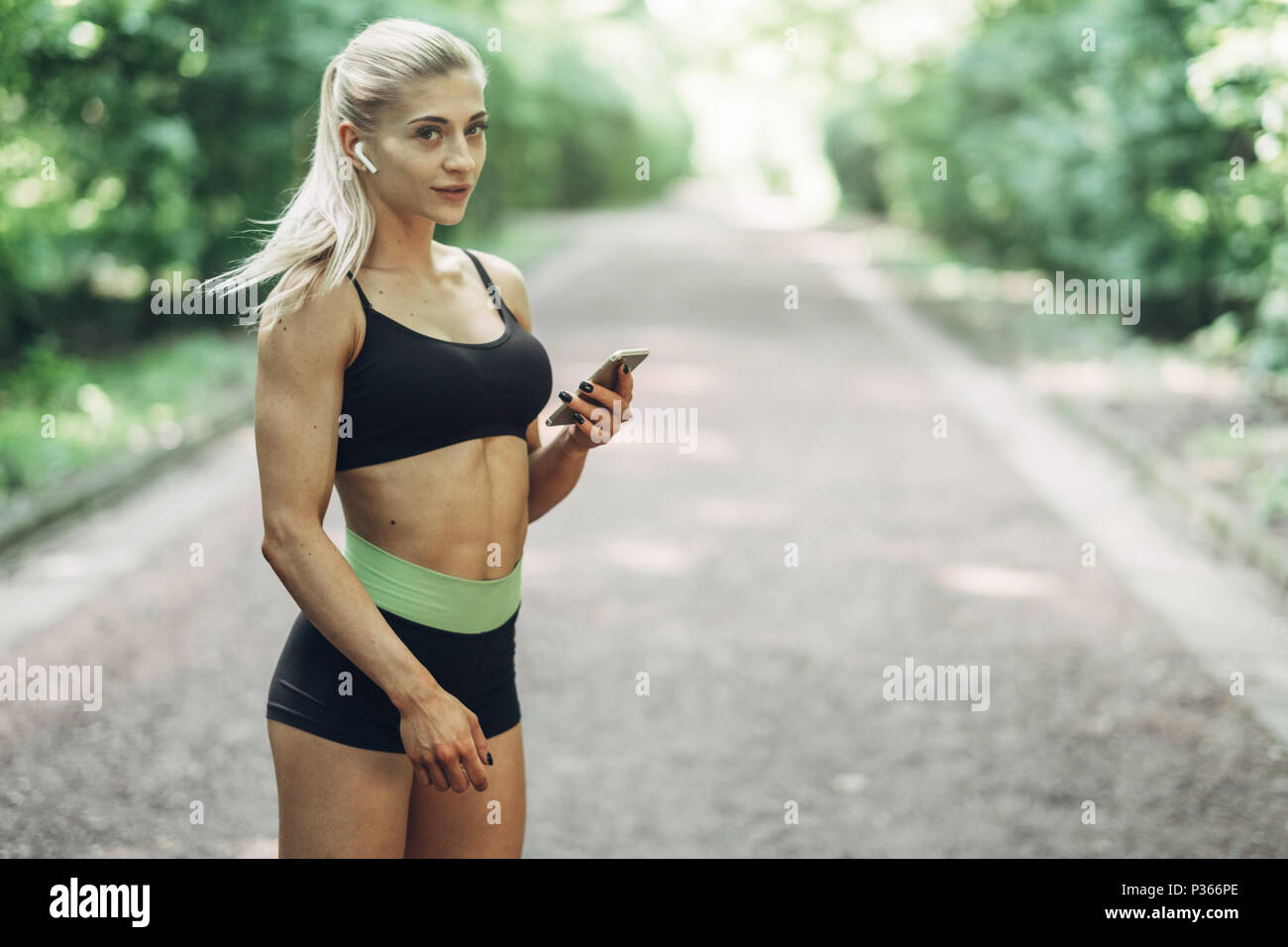Runner donna nella mattinata estiva Park ascoltare musica sullo smartphone utilizzando gli auricolari Bluetooth. Femmina ragazza Fitness jogging sul percorso esterno. Foto Stock
