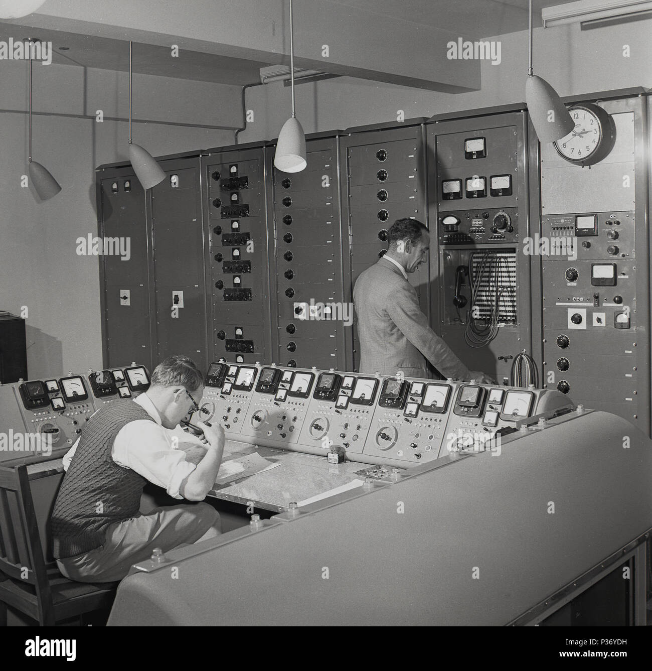 Anni '1950, un operatore wireless Marconi, con tubo seduto a un banco di controllo, circondato da una banca di numerosi quadranti e interruttori, Inghilterra, Regno Unito. Un altro tecnico che si trova in una fila di armadi metallici, davanti a un telefono e a numerosi cavi. Fondata da Guglielmo Marconi nel 1897, la società Marconi era un nome di spicco nel settore delle comunicazioni radio e a lunga distanza. Foto Stock
