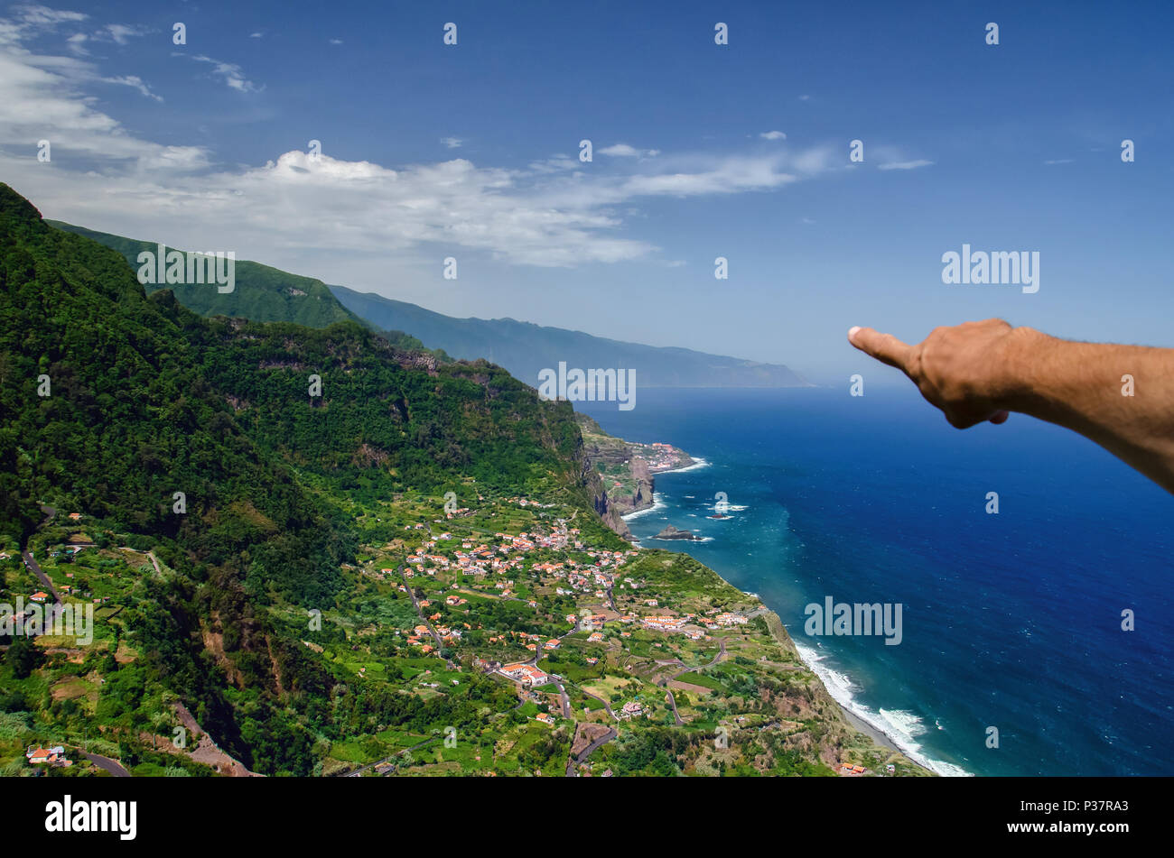 Mano del maschio di puntamento turistica in vista della costa nord dell'isola di Madeira, Santana città circondata da alte montagne e profondo blu oceano Atlantico. Laborato Foto Stock