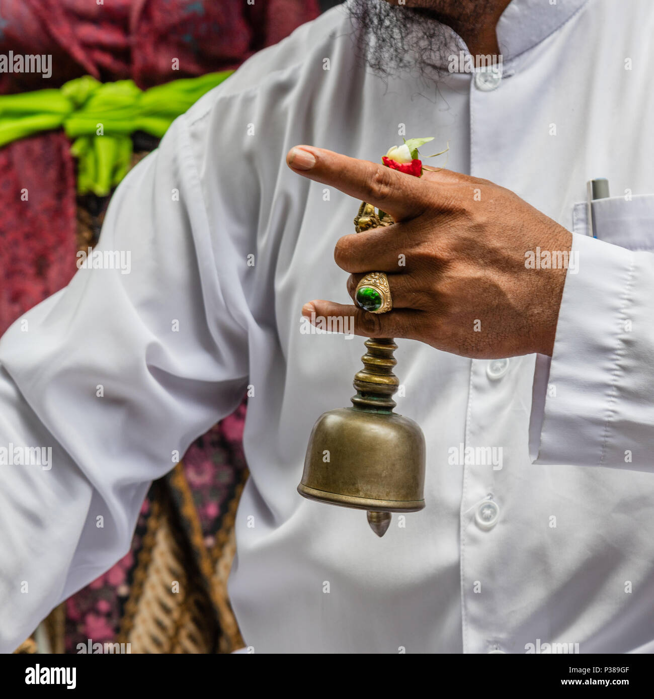 Balinese sacerdote Indù pemangku vestite di bianco tenendo un bell (Bidschel) durante la famiglia tiga bulanan cerimonia. Bali, Indonesia. Immagine quadrata Foto Stock