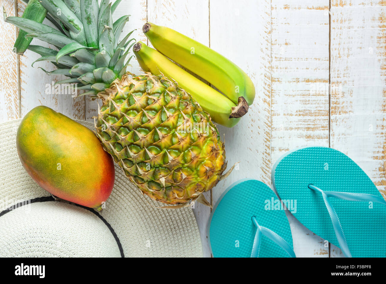 Piatto disposizione laici composizione di frutti tropicali ananas Banane Mango donne Hat pantofole blu su bianco Planked Sfondo legno. Vacanza in viaggio Foto Stock