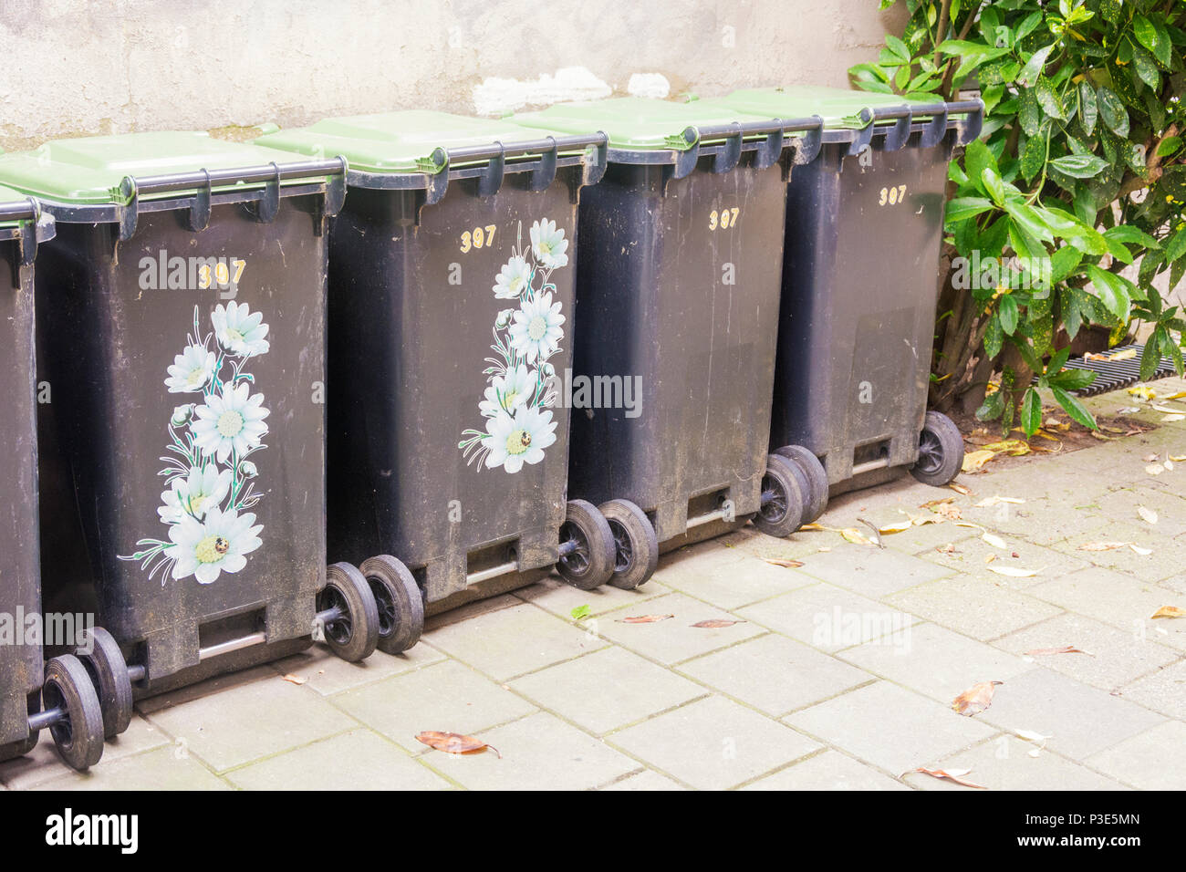 Fila di contenitori per rifiuti, alcuni decorati per renderle bellissime, nella città contro un vecchio muro di cemento. Una pianta verde accanto ai contenitori come na Foto Stock